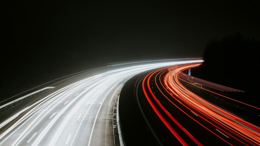 Langsame Lichtspuren von Fahrzeugen auf einer dunklen Straße, mit baumbewachsenen Ufern.
