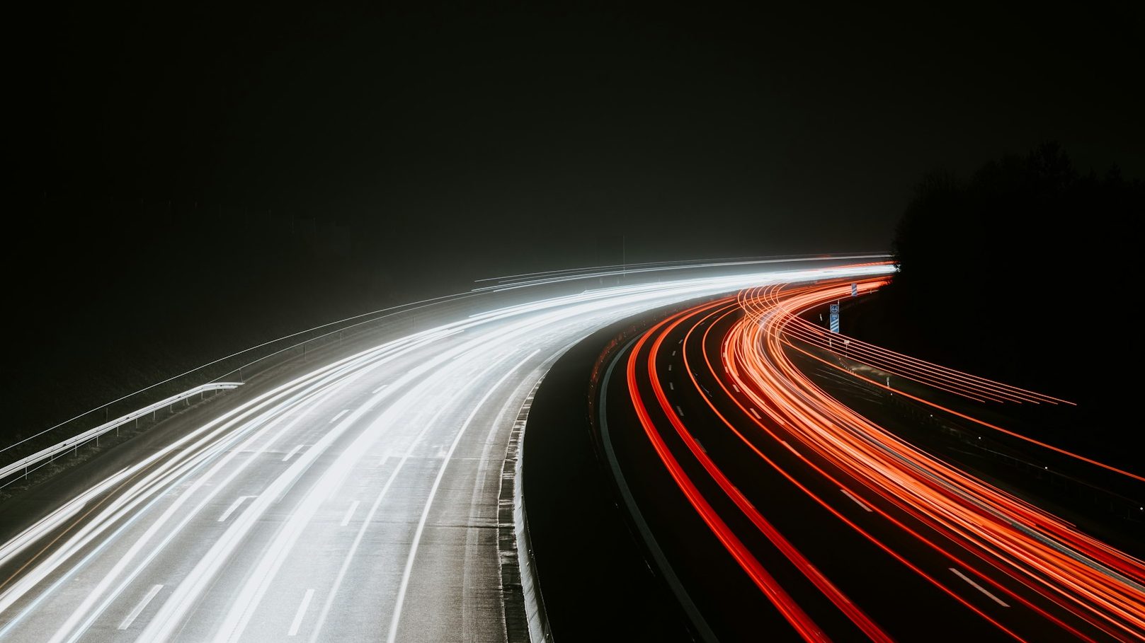 Langsame Lichtspuren von Fahrzeugen auf einer dunklen Straße, mit baumbewachsenen Ufern.