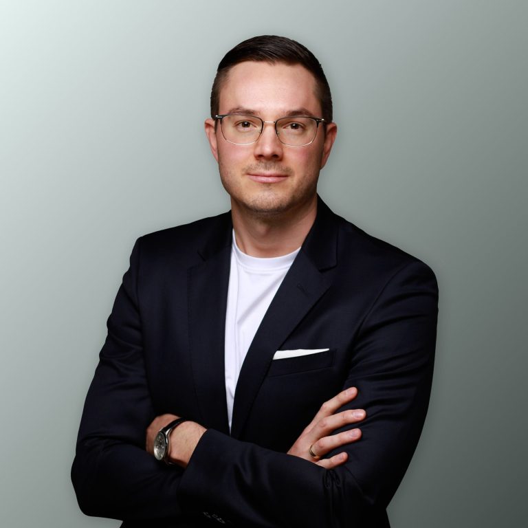 Mann in Anzug mit überkreuzten Armen vor einem neutralen Hintergrund.