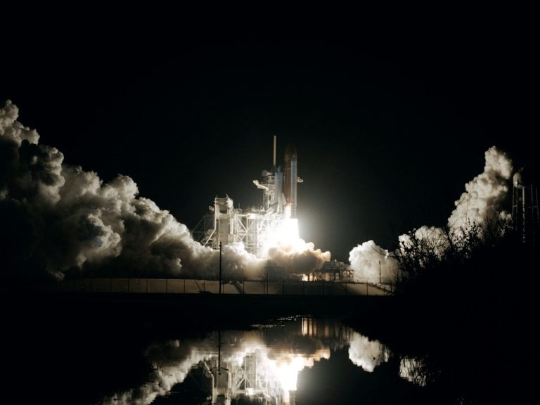 Rakete startet bei Nacht, umgeben von Rauch, mit Reflexion in ruhigem Wasser.